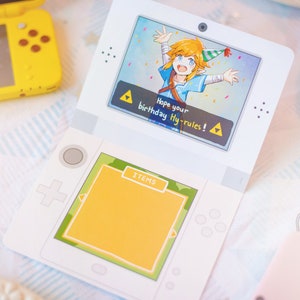 Puede incluir: Un recorte de papel blanco con forma de consola de juegos port&aacute;til. La pantalla superior muestra un personaje de dibujos animados con un gorro de fiesta y el texto 'Hope your birthday Hy-rules!'. La pantalla inferior tiene un cuadrado amarillo 'ITEMS'.