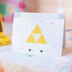 Puede incluir: Una tarjeta rectangular blanca con un emblema central de tri&aacute;ngulo amarillo compuesto por tres tri&aacute;ngulos m&aacute;s peque&ntilde;os. La tarjeta est&aacute; decorada con un patr&oacute;n de peque&ntilde;as chispas multicolores en rosa, amarillo y azul, e incluye dos iconos circulares oscuros.