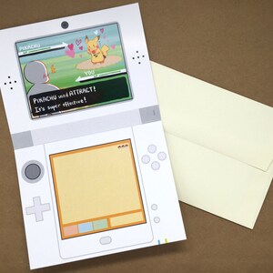 Puede incluir: Una tarjeta de felicitaci&oacute;n blanca dise&ntilde;ada para parecer una consola de juegos port&aacute;til. La pantalla superior muestra una escena de batalla con Pikachu y el texto PIKACHU used ATTRACT! It's super effective! La pantalla inferior est&aacute; vac&iacute;a. Incluye un sobre color crema.