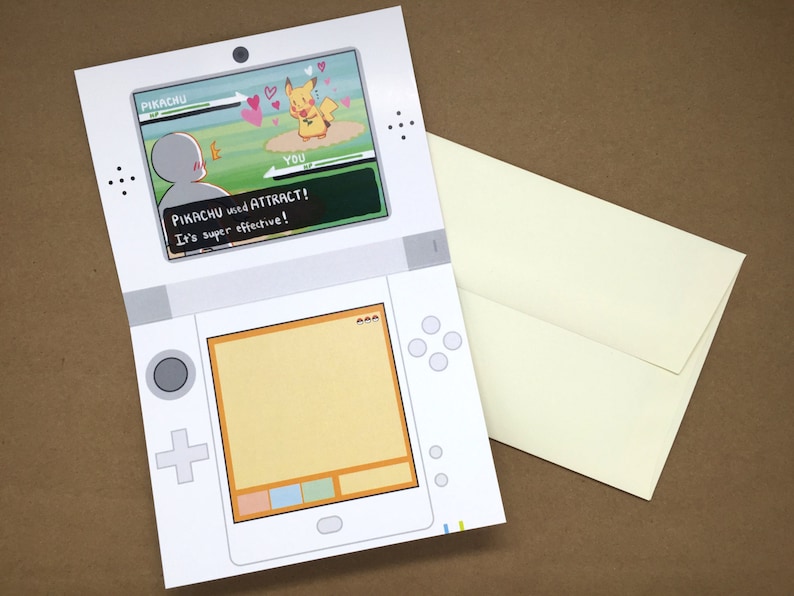 Puede incluir: Una tarjeta de felicitaci&oacute;n blanca dise&ntilde;ada para parecer una consola de juegos port&aacute;til. La pantalla superior muestra una escena de batalla con Pikachu y el texto PIKACHU used ATTRACT! It's super effective! La pantalla inferior est&aacute; vac&iacute;a. Incluye un sobre color crema.