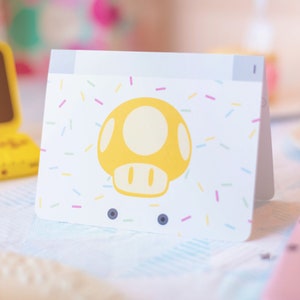 Puede incluir: Una tarjeta de felicitaci&oacute;n rectangular blanca con un gr&aacute;fico de hongo amarillo centrado en la parte frontal. El dise&ntilde;o de la tarjeta incluye un patr&oacute;n de peque&ntilde;as chispas multicolores esparcidas sobre el fondo blanco. La tarjeta est&aacute; doblada.