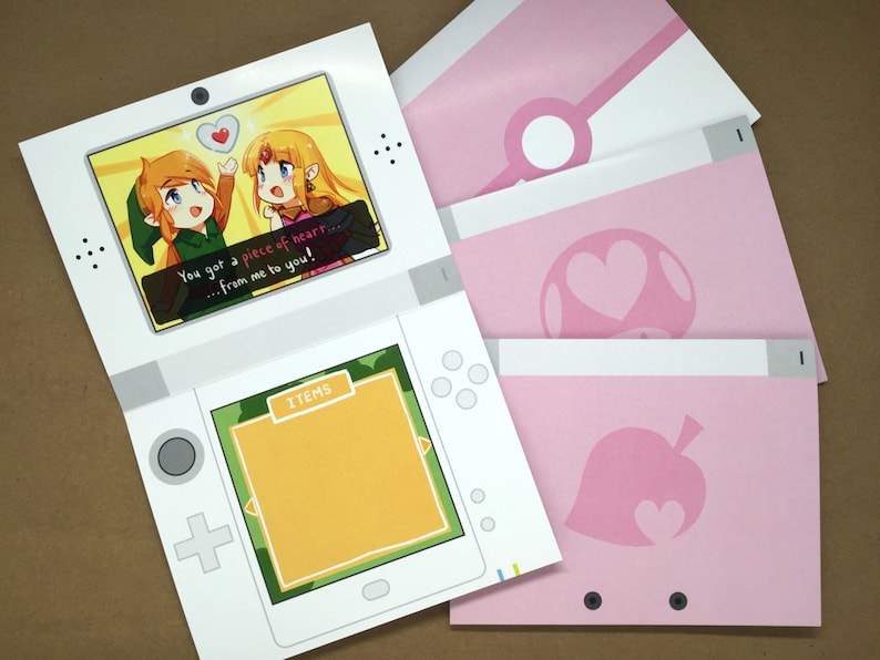 Puede incluir: Un recorte de papel con forma de consola de juegos port&aacute;til que muestra una pantalla con dos personajes y el texto You got a piece of heart... from me to you! y un men&uacute; de objetos, junto a tres insertos de papel rosa con varios iconos.