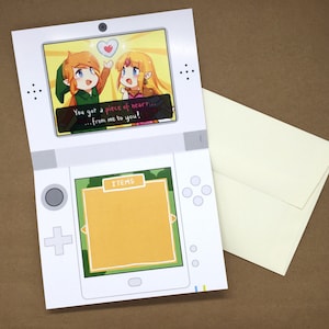 Puede incluir: Una tarjeta de felicitaci&oacute;n blanca con forma de consola de juegos port&aacute;til. La pantalla superior muestra una ilustraci&oacute;n de dos personajes con el texto, You got a piece of heart... from me to you! La pantalla inferior muestra un cuadro amarillo vac&iacute;o.