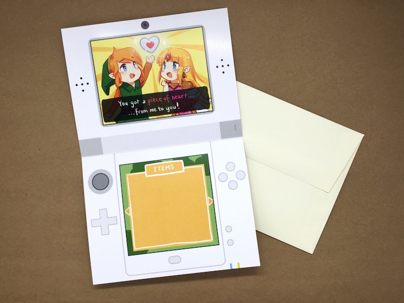 Puede incluir: Una tarjeta de felicitaci&oacute;n blanca con forma de consola de juegos port&aacute;til. La pantalla superior muestra una ilustraci&oacute;n de dos personajes con el texto, You got a piece of heart... from me to you! La pantalla inferior muestra un cuadro amarillo vac&iacute;o.