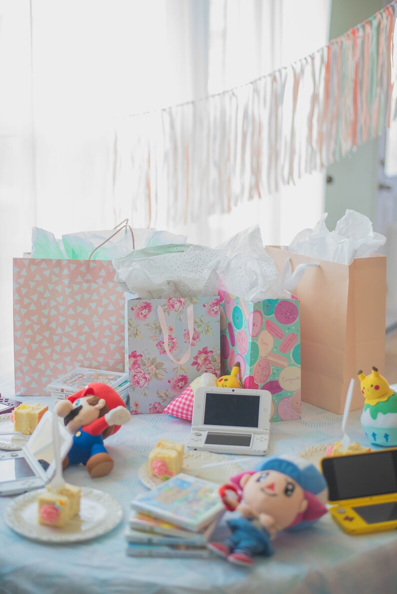 Puede incluir: Una mesa de fiesta con bolsas de regalo con patrones florales, de macarons y geom&eacute;tricos, una consola de juegos port&aacute;til blanca, una consola amarilla, peluches de Mario y un aldeano, una figura de Pikachu y pasteles peque&ntilde;os en platos.