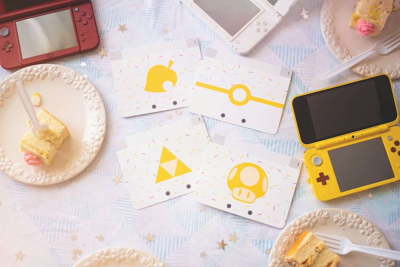 Puede incluir: Cuatro tarjetas rectangulares blancas con iconos amarillos de una hoja, un c&iacute;rculo, un tri&aacute;ngulo y un champi&ntilde;&oacute;n, decoradas con virutas de colores. Las tarjetas est&aacute;n dispuestas entre tres consolas de juegos port&aacute;tiles y platos de pastel.
