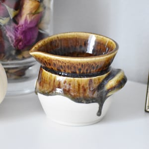 May include: A small, white ceramic creamer with a brown glaze. The glaze has a drip effect and is unevenly applied, creating a rustic look.