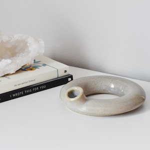 Puede incluir: Un anillo de cerámica gris claro con un pequeño agujero en un lado. El anillo está sobre una superficie blanca con dos libros y un cristal blanco.