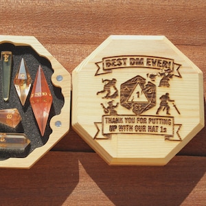 May include: An octagonal wooden box with a removable lid featuring laser-engraved text reading BEST DM EVER! THANK YOU FOR PUTTING UP WITH OUR NAT 1s and a central d20 graphic. The box contains seven translucent amber-coloured polyhedral dice.