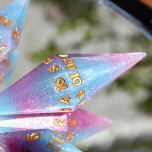 Puede incluir: Un juego de dados poli&eacute;dricos con un acabado de resina transl&uacute;cida en tonos azul y rosa con part&iacute;culas de purpurina fina. Cada dado presenta bordes geom&eacute;tricos afilados y n&uacute;meros grabados rellenos de un pigmento dorado met&aacute;lico.