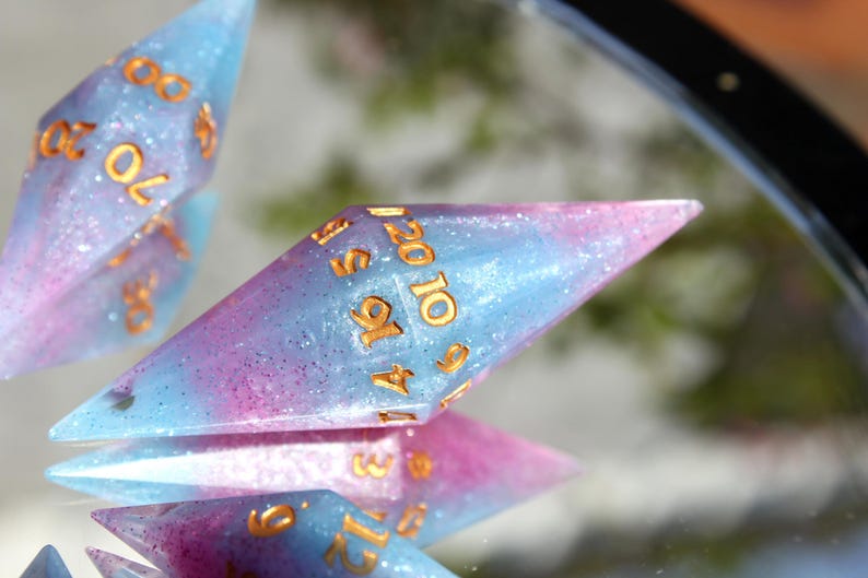 Puede incluir: Un juego de dados poli&eacute;dricos con un acabado de resina transl&uacute;cida en tonos azul y rosa con part&iacute;culas de purpurina fina. Cada dado presenta bordes geom&eacute;tricos afilados y n&uacute;meros grabados rellenos de un pigmento dorado met&aacute;lico.