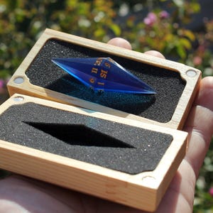 Puede incluir: Un dado de juego azul en forma de diamante con números dorados, presentado en una caja de madera abierta con acolchado de espuma negra. La caja es rectangular con un acabado de madera natural. El dado se sostiene en la mano de una persona.