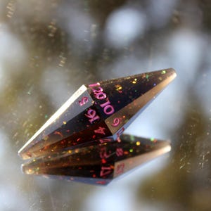 Op de afbeelding: Een zwarte, langwerpige, veelzijdige dobbelsteen met roze cijfers en kleurrijke glitterinsluitsels. De dobbelsteen wordt weerspiegeld in een spiegelend oppervlak, wat de geometrische vorm en het ontwerp laat zien. Dit item is voor tabletop gaming.