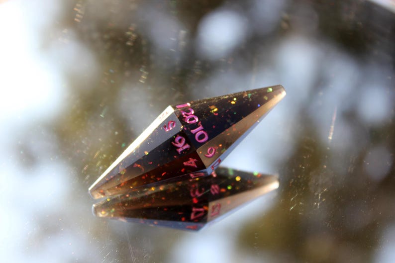 Op de afbeelding: Een zwarte, langwerpige, veelzijdige dobbelsteen met roze cijfers en kleurrijke glitterinsluitsels. De dobbelsteen wordt weerspiegeld in een spiegelend oppervlak, wat de geometrische vorm en het ontwerp laat zien. Dit item is voor tabletop gaming.
