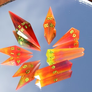 May include: A collection of gemstone dice in shades of red, orange, and yellow, with gold-colored numbers. The dice are arranged in a starburst pattern, reflecting on a mirrored surface. The dice are designed for tabletop gaming.