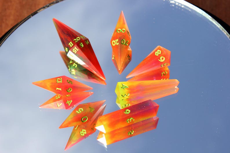 May include: A collection of gemstone dice in shades of red, orange, and yellow, with gold-colored numbers. The dice are arranged in a starburst pattern, reflecting on a mirrored surface. The dice are designed for tabletop gaming.