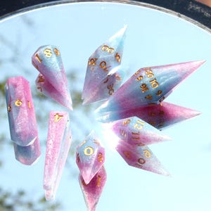 Puede incluir: Un juego de dados poli&eacute;dricos que presenta una mezcla de resina en azul claro y rosa con un acabado de purpurina sutil. Cada dado tiene puntas afiladas y alargadas en forma de cristal, marcadas con n&uacute;meros dorados de f&aacute;cil lectura.