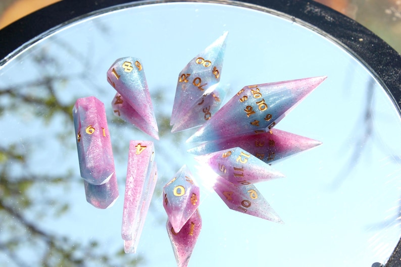 Puede incluir: Un juego de dados poli&eacute;dricos que presenta una mezcla de resina en azul claro y rosa con un acabado de purpurina sutil. Cada dado tiene puntas afiladas y alargadas en forma de cristal, marcadas con n&uacute;meros dorados de f&aacute;cil lectura.