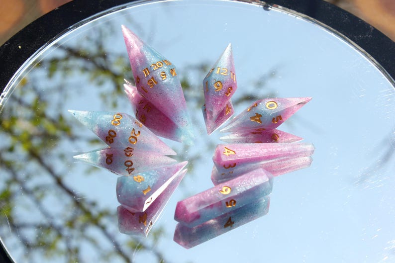 Puede incluir: Un conjunto de dados poli&eacute;dricos con una mezcla de resina rosa y azul con purpurina incrustada. Cada dado tiene puntas afiladas y alargadas, marcadas con n&uacute;meros de color dorado, dispuestos en un patr&oacute;n circular sobre una superficie reflectante.