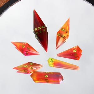 May include: A collection of seven polyhedral dice in various shapes, arranged in a circular pattern. The dice are translucent with a gradient of red, orange, and yellow hues. Each die features gold-colored numbers.