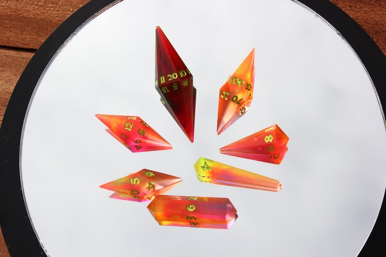 May include: A collection of seven polyhedral dice in various shapes, arranged in a circular pattern. The dice are translucent with a gradient of red, orange, and yellow hues. Each die features gold-colored numbers.