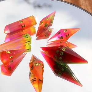 May include: A collection of colorful, gem-like polyhedral dice. The dice are a mix of red, orange, and yellow with gold numbering. The dice are arranged in a circular pattern on a reflective surface, creating a unique display.