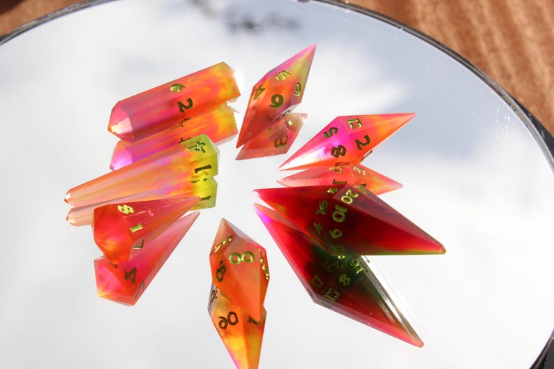 May include: A collection of colorful, gem-like polyhedral dice. The dice are a mix of red, orange, and yellow with gold numbering. The dice are arranged in a circular pattern on a reflective surface, creating a unique display.