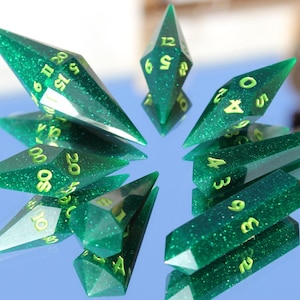 May include: A set of green, glittery polyhedral dice with yellow numbering. The dice are arranged in a circular pattern, reflecting on a shiny surface. The set includes various shapes for role-playing games.