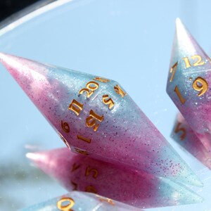 Puede incluir: Dos dados poli&eacute;dricos alargados en forma de diamante, hechos de resina transl&uacute;cida con un degradado de rosa y azul claro e infundidos con purpurina fina. Los dados tienen n&uacute;meros dorados grabados, incluyendo un 20 visible en el dado mayor.