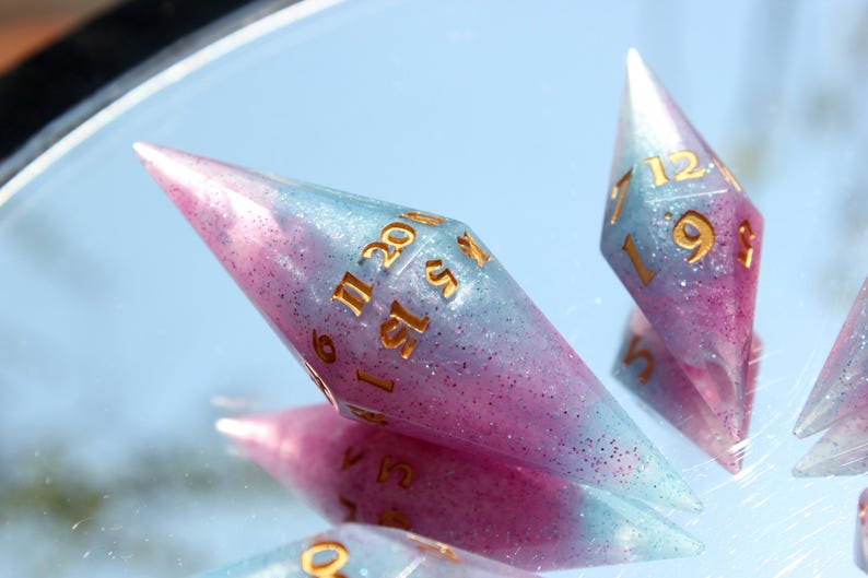 Puede incluir: Dos dados poli&eacute;dricos alargados en forma de diamante, hechos de resina transl&uacute;cida con un degradado de rosa y azul claro e infundidos con purpurina fina. Los dados tienen n&uacute;meros dorados grabados, incluyendo un 20 visible en el dado mayor.