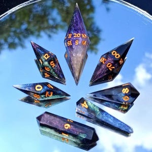 May include: A set of seven polyhedral dice, each with a unique, pointed shape. The dice are a mix of dark blue, purple, and gold, with gold numbering. The dice are arranged in a circle on a reflective surface.