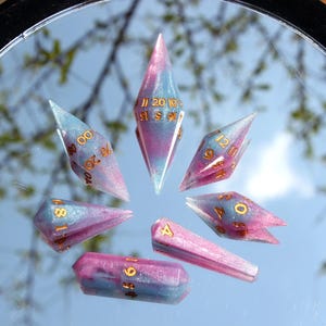 Puede incluir: Un juego de siete dados poli&eacute;dricos con un acabado de resina en espiral rosa y azul y n&uacute;meros de color dorado. Los dados est&aacute;n dispuestos en un patr&oacute;n circular sobre una superficie reflectante, con formas de diamante alargadas y puntiagudas.
