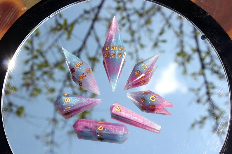 Puede incluir: Un juego de siete dados poli&eacute;dricos con un acabado de resina en espiral rosa y azul y n&uacute;meros de color dorado. Los dados est&aacute;n dispuestos en un patr&oacute;n circular sobre una superficie reflectante, con formas de diamante alargadas y puntiagudas.