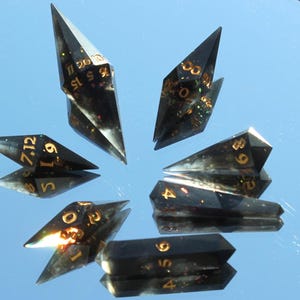 Könnte beinhalten: Ein Satz von sieben schwarzen und goldenen polyedrischen Würfeln, jeder mit einer einzigartigen geometrischen Form. Die Würfel bestehen aus einem durchscheinenden Material mit goldfarbenen Zahlen und Glitzerakzenten. Sie sind auf einer reflektierenden Oberfläche angeordnet.