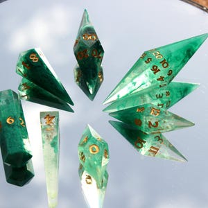 May include: A collection of green and gold resin dice, including D4, D6, D8, D10, D12, and D20 dice. The dice are arranged on a reflective surface, showcasing their geometric shapes and gold numbering. The dice are for tabletop games.