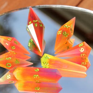 May include: A collection of colorful, gem-like polyhedral dice. The dice are primarily orange and red with green numbering. The dice are arranged on a reflective surface, creating a mirrored effect. The dice are designed for tabletop gaming.