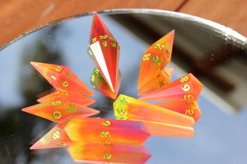 May include: A collection of colorful, gem-like polyhedral dice. The dice are primarily orange and red with green numbering. The dice are arranged on a reflective surface, creating a mirrored effect. The dice are designed for tabletop gaming.