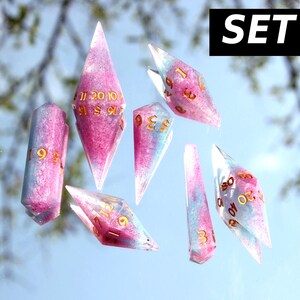 Puede incluir: Un juego de siete dados poli&eacute;dricos con una mezcla de resina rosa y azul claro con n&uacute;meros de color dorado. Los dados tienen formas alargadas de bordes afilados y est&aacute;n dispuestos sobre una superficie reflectante. El texto SET #5 es visible.
