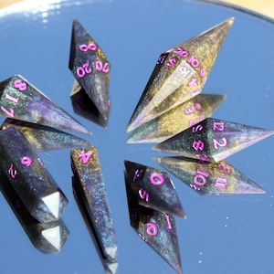 Puede incluir: Un conjunto de dados de múltiples caras con un acabado oscuro e iridiscente y numeración rosa. Los dados están dispuestos sobre una superficie reflectante, creando un efecto de espejo. Los dados están diseñados para juegos de mesa.