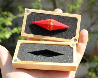 Puede incluir: Un dado rojo de múltiples caras con números dorados, colocado en una caja de madera abierta con inserciones de espuma negra. La caja se sostiene en una mano, con un fondo verde borroso.