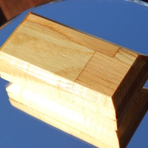 Op de afbeelding: Een rechthoekige houten kist met een lichtbruine afwerking. De kist is gemaakt van meerdere stukken hout die aan elkaar zijn gezet, met een facetgeslepen bovenkant. De kist wordt weerspiegeld in een spiegelend oppervlak.