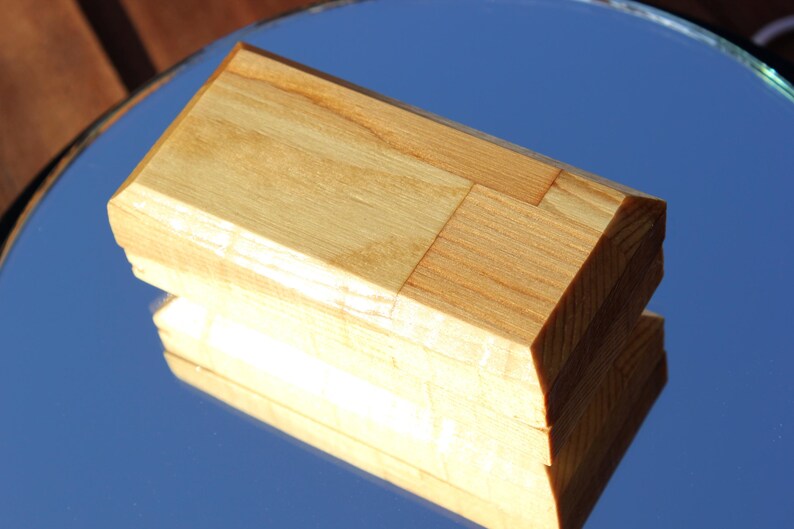 Op de afbeelding: Een rechthoekige houten kist met een lichtbruine afwerking. De kist is gemaakt van meerdere stukken hout die aan elkaar zijn gezet, met een facetgeslepen bovenkant. De kist wordt weerspiegeld in een spiegelend oppervlak.
