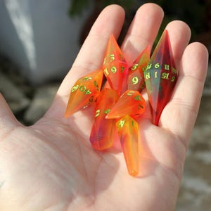 May include: A collection of translucent, orange and red polyhedral dice with green numbering, held in a hand. The dice are elongated and pointed, with a variety of numbers visible. The dice are for tabletop gaming.