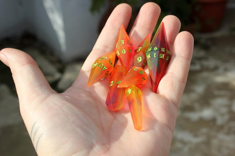 May include: A collection of translucent, orange and red polyhedral dice with green numbering, held in a hand. The dice are elongated and pointed, with a variety of numbers visible. The dice are for tabletop gaming.