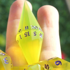 以下が含まれることがあります： 銀色の数字が入った、半透明の黄色の多面体サイコロのコレクション。1つのサイコロが手に持たれ、他のサイコロが下に散らばっています。サイコロはテーブルトップRPG用に設計されています。