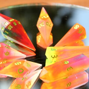 May include: A collection of colorful, gemstone-like polyhedral dice. The dice are a mix of orange, yellow, pink, and red with green numbers. The dice are arranged on a reflective surface.