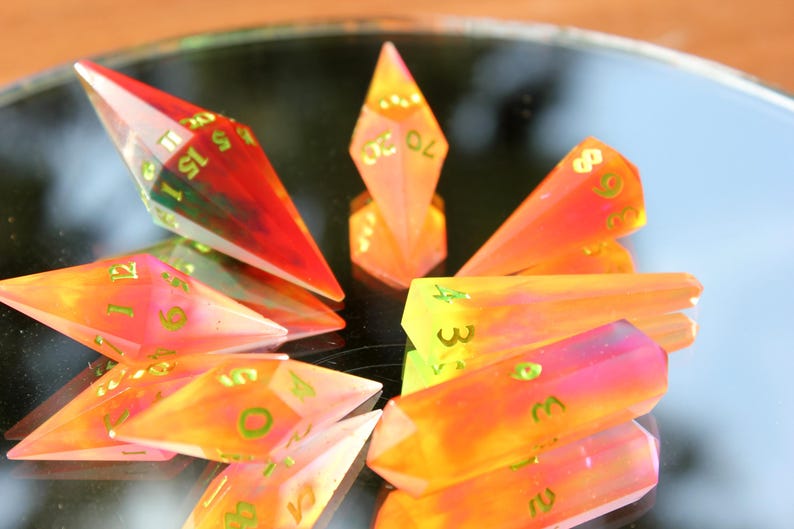 May include: A collection of colorful, gemstone-like polyhedral dice. The dice are a mix of orange, yellow, pink, and red with green numbers. The dice are arranged on a reflective surface.