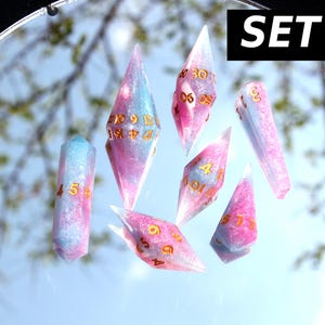 Puede incluir: Un conjunto de siete dados poli&eacute;dricos etiquetados como SET #4, que presentan una mezcla de resina transl&uacute;cida rosa y azul claro con purpurina incrustada. Cada dado tiene n&uacute;meros de color dorado grabados en sus caras geom&eacute;tricas.