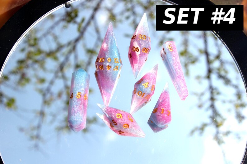 Puede incluir: Un conjunto de siete dados poli&eacute;dricos etiquetados como SET #4, que presentan una mezcla de resina transl&uacute;cida rosa y azul claro con purpurina incrustada. Cada dado tiene n&uacute;meros de color dorado grabados en sus caras geom&eacute;tricas.