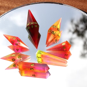 May include: A collection of colorful, geometric dice with gold numbering, reflecting on a mirrored surface. The dice are primarily red, orange, and yellow, with a few in shades of pink and purple. The dice are arranged in a circular pattern.